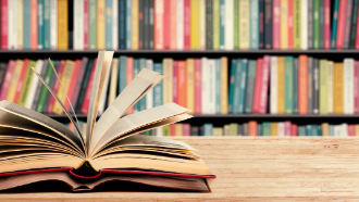 Picture of a book opened up and fanning on a table with a shelf of books in the background